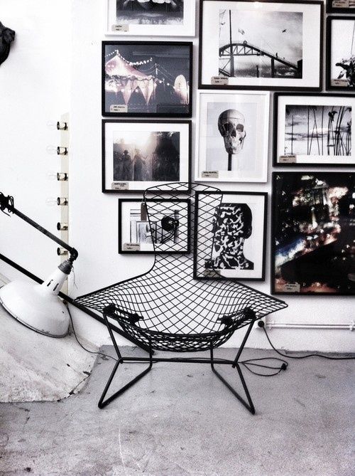 A modern, black wireframe chair is placed in front of a gallery wall filled with black and white framed artwork, including images of skulls and abstract designs. The industrial lamp beside the chair adds to the edgy, rock and roll atmosphere of the space.
