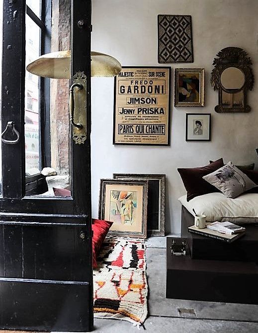 An eclectic living space with a mix of vintage and modern decor. The room features a black-framed glass door, a gallery wall with framed posters and artwork, and a cozy seating area with plush cushions and layered rugs, blending dark and light elements.