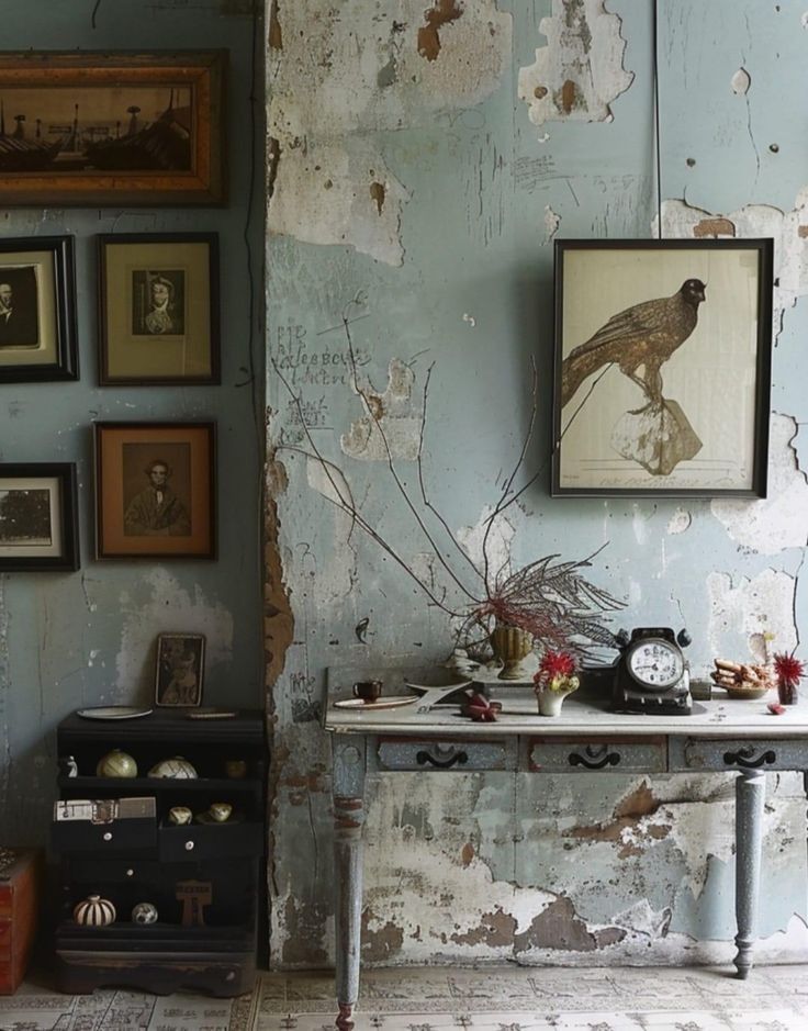 A rustic interior with a distressed blue wall, adorned with framed botanical prints and vintage photographs. A wooden console table is decorated with an antique clock, dried botanical arrangements, and small decorative items.
