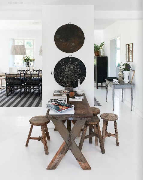 A dining area with a rustic wooden table surrounded by wooden stools, with black and white wall decor and a view into a living room with a black and white striped rug.