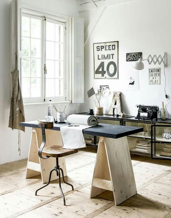 A bright and airy home workspace with a modern sawhorse desk, a wooden swivel chair, and decor elements like a typewriter and vintage signs. The room is filled with natural light from a large window, adding warmth to the wooden floor and white walls.