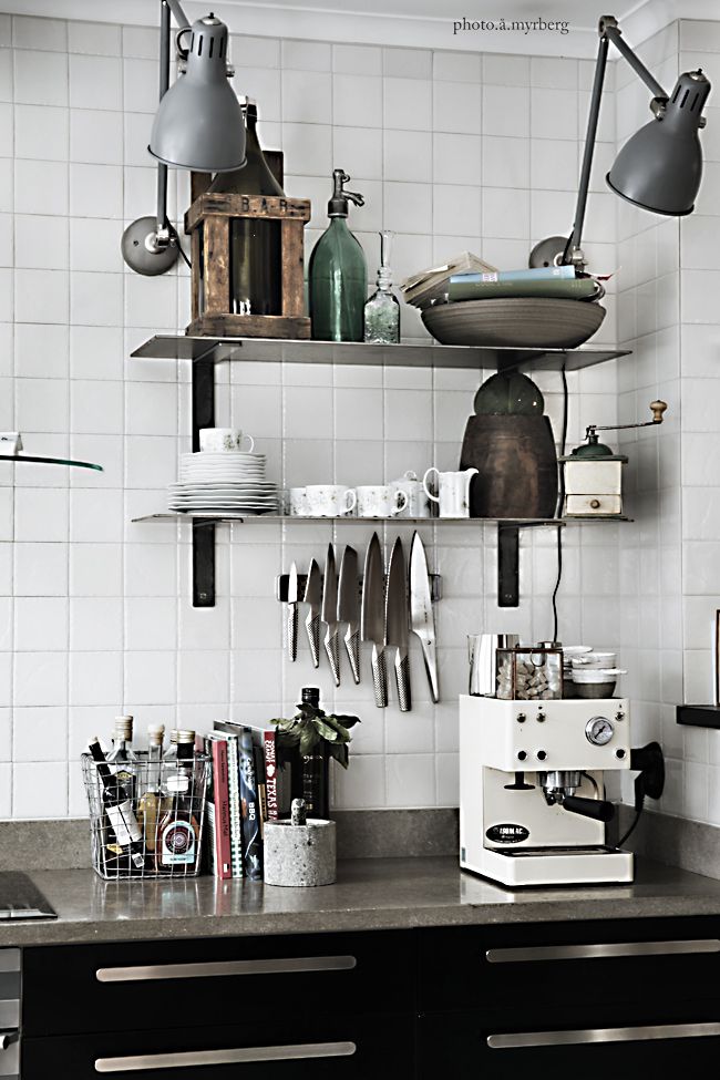 Modern eclectic kitchen with open shelves featuring a mix of vintage and contemporary elements, including a green glass bottle, a wooden crate, and a collection of white crockery, with hanging knives and a coffee machine on the countertop below.