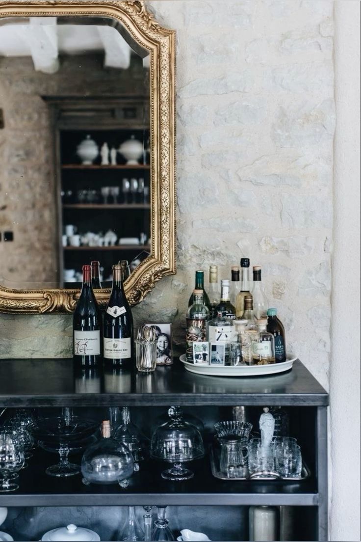 A stylish bar cart setup against a stone wall with a large ornate gold-framed mirror. The cart is adorned with bottles of wine, spirits, and glassware, showcasing an elegant and eclectic mix of vintage and contemporary decor.