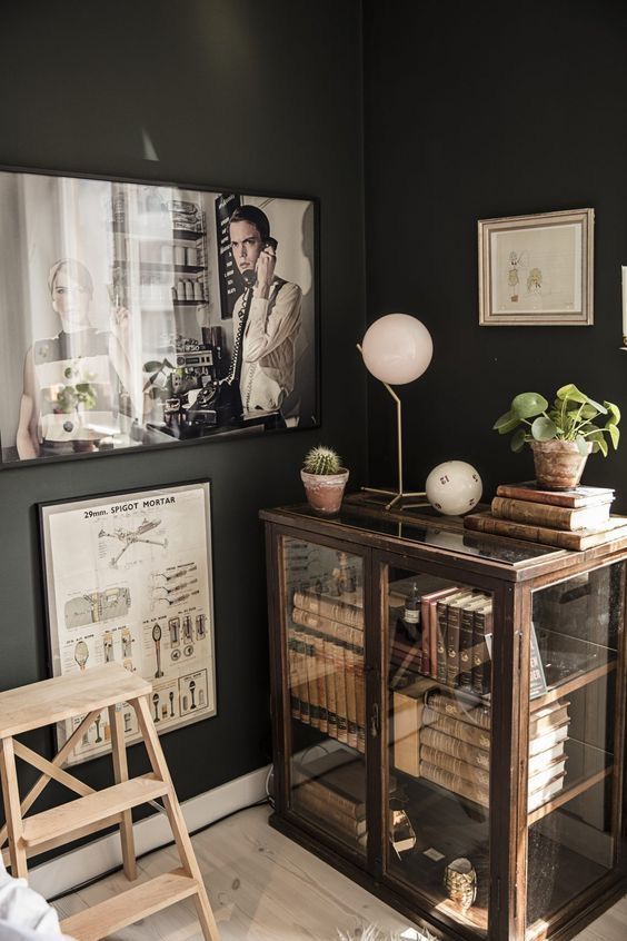 A cozy corner with a vintage wooden cabinet filled with books, topped with decorative items like a lamp, potted plants, and vintage books. The dark green walls are decorated with framed artwork, adding a warm and sophisticated ambiance to the room.