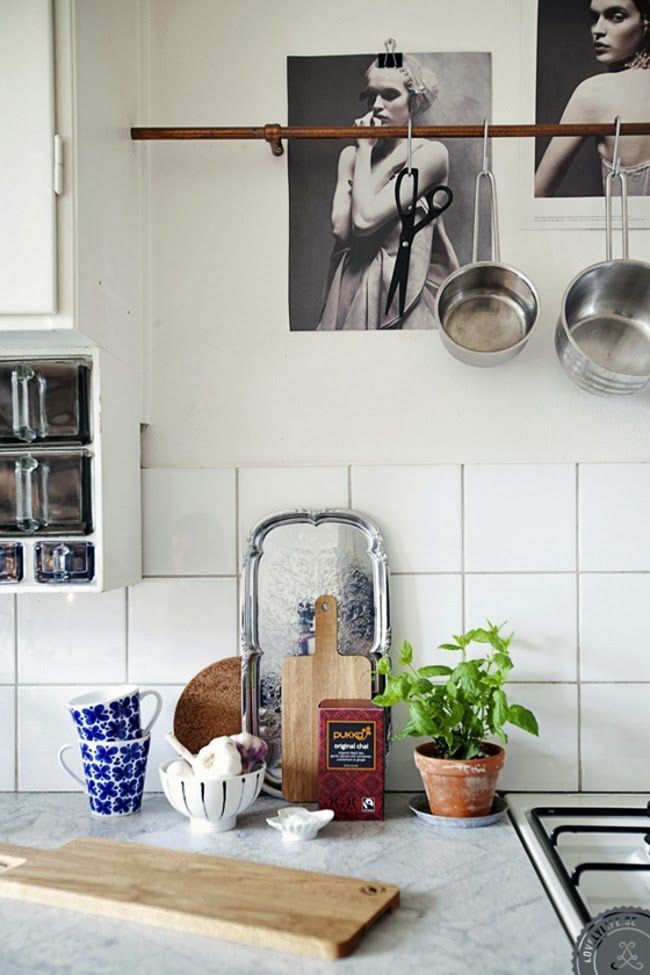 Small eclectic kitchen corner with a rustic wooden rod used for hanging pots, pans, and scissors, a minimalist display of cutting boards, blue patterned cups, and a potted herb plant on the countertop.