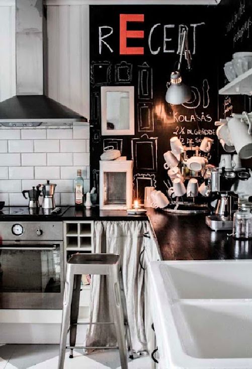 An eclectic kitchen corner with a black chalkboard wall covered in various drawings and recipes. The space includes a stainless steel oven, open shelving with kitchen tools, a sink with a white backsplash, and a metal stool. The word ‘RECET’ is prominently written in red and white chalk