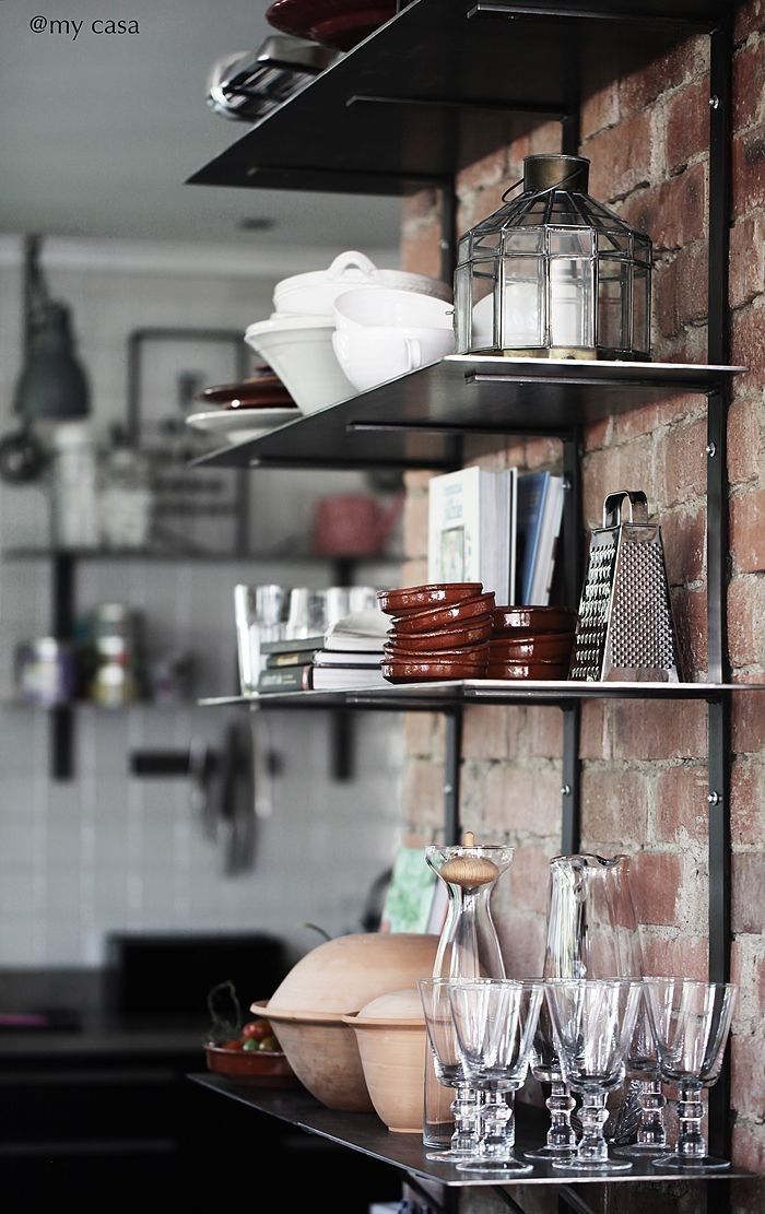 Eclectic kitchen shelving with black metal shelves mounted on a brick wall, displaying white ceramic dishes, clear glassware, a metal lantern, and various kitchen items in a layered, visually interesting arrangement.