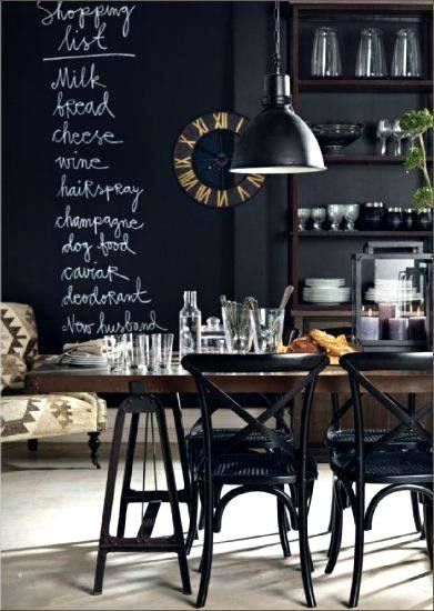 A cozy dining area with a chalkboard wall displaying a handwritten shopping list. The room features dark wood furniture, a black metal pendant light, and a mix of modern and vintage decor elements, including a large wall clock and glassware on a wooden dining table.