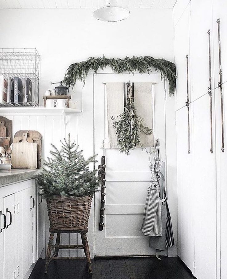 A cozy white kitchen with a small Christmas tree in a woven basket, complemented by holiday greenery above the door and rustic wooden accents.