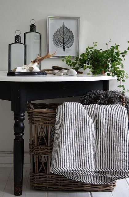 A half-circle black accent table with a white top stands against a light wall. It is adorned with natural decor, including a potted plant, lanterns, and seashells. Below the table, a woven basket holds striped blankets, enhancing the rustic and cozy aesthetic of the space.