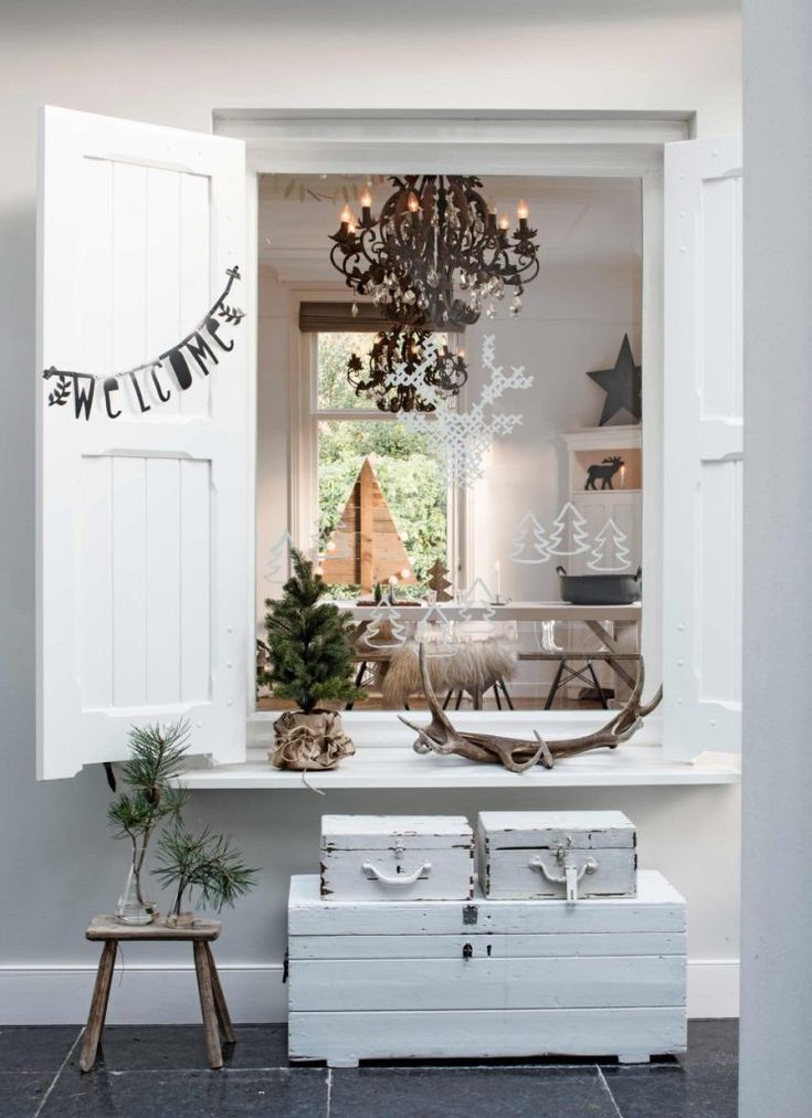 A Scandinavian-inspired interior with white and natural accents, decorated for the winter season. Pine branches in vases, antlers, and simple wooden crates create a welcoming, understated holiday look. A chandelier and frosted window decals add a touch of elegance to the rustic decor.