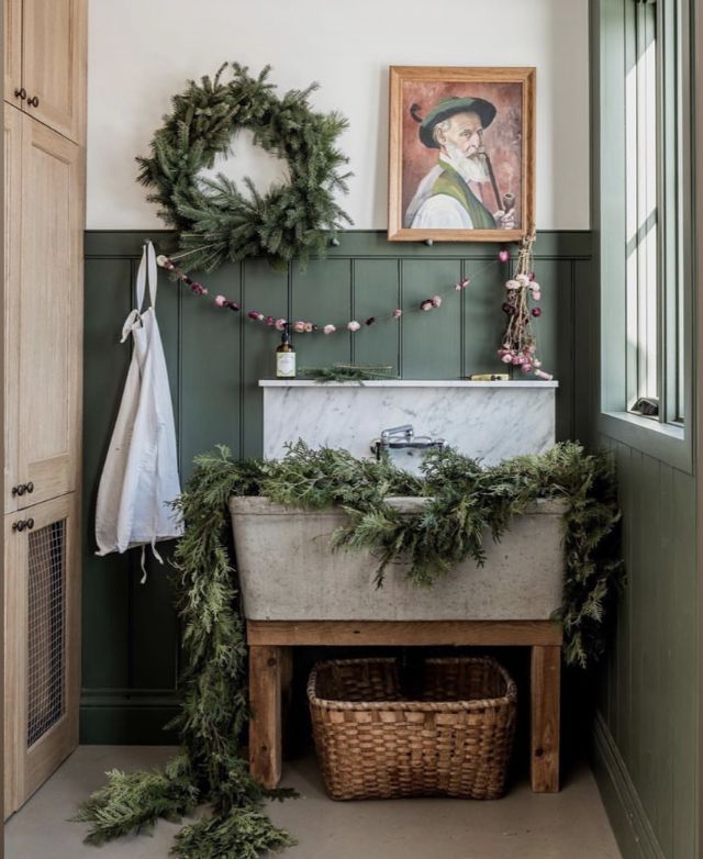A charming green-walled bathroom featuring a vintage-style sink adorned with pine garlands, a wicker basket below, and a wreath on the wall, enhancing the room’s rustic, festive feel.