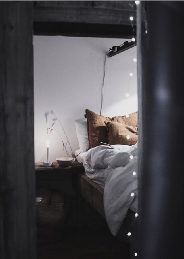 Cozy Scandinavian bedroom with warm brown cushions, soft white bedding, and string fairy lights. A candle glows softly on the bedside table, adding warmth and serenity to the room.