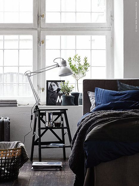 A modern bedroom with a mix of deep blue bedding and monochromatic decor. A black side table with a desk lamp sits near a large window, adding light and greenery to the dark, cozy space.