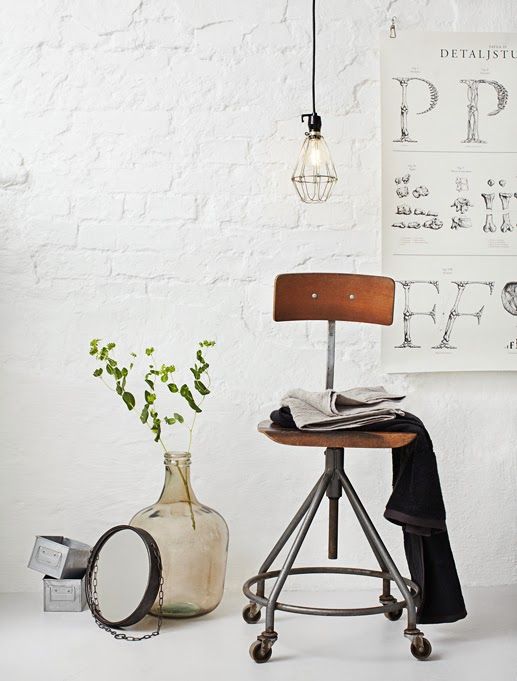A cozy setup against a white brick wall featuring an industrial-style stool with a wooden seat, a wire pendant light hanging from the ceiling, and a large glass bottle used as decor. The space includes vintage artwork on the wall, creating a trendy mix of industrial and rustic design.