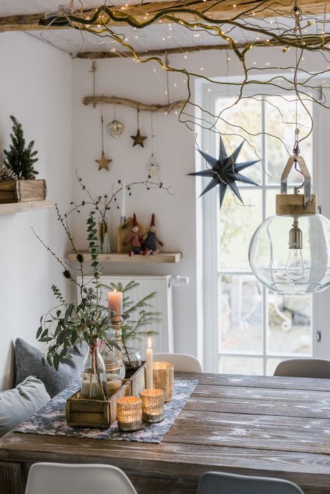 A cozy dining room decorated with natural branches and soft white string lights hanging from the ceiling, creating a warm and rustic ambiance. The table is set with candles, greenery in glass vases, and a few simple Christmas ornaments, providing a subtle transition from fall to winter decor.