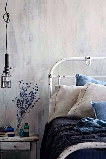 A vintage white metal bedframe with pillows in shades of blue and beige. A hanging industrial light and blue flowers in a glass vase on a bedside table add rustic charm to the room’s soft, serene blue decor.