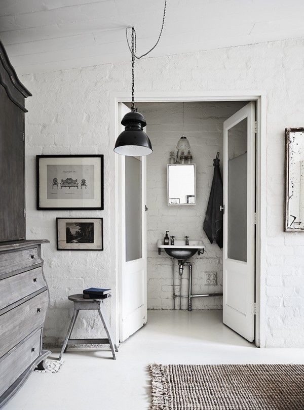A stylish room featuring a white brick wall with a minimalist sink area visible through an open door. The room is accented with black pendant lighting, framed artwork, and a vintage stool next to a dark wooden dresser. The space combines rustic charm with modern industrial elements.