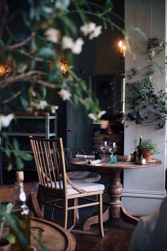 A charming Scandinavian-inspired dining nook with a wooden table and simple wooden chairs. The space is softened by natural greenery, a candle-lit ambiance, and warm lighting from the wall sconces.