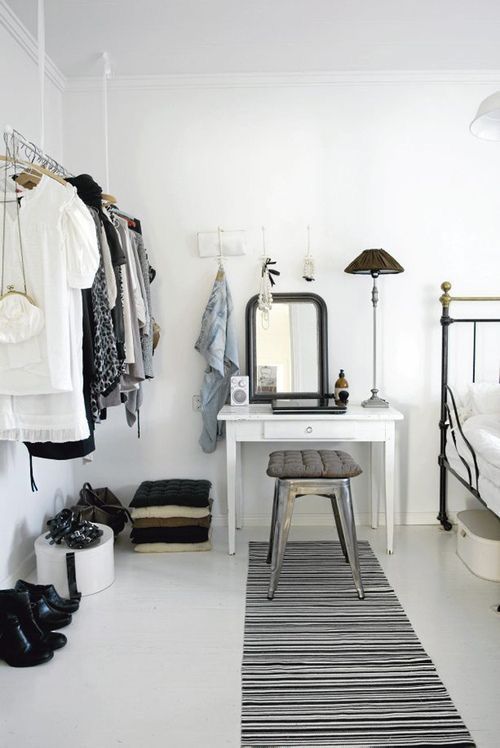 A minimalist white bedroom with an open clothing rack, a white vanity, and a black-and-white striped rug. The decor combines industrial and modern elements, creating a clean, airy aesthetic.