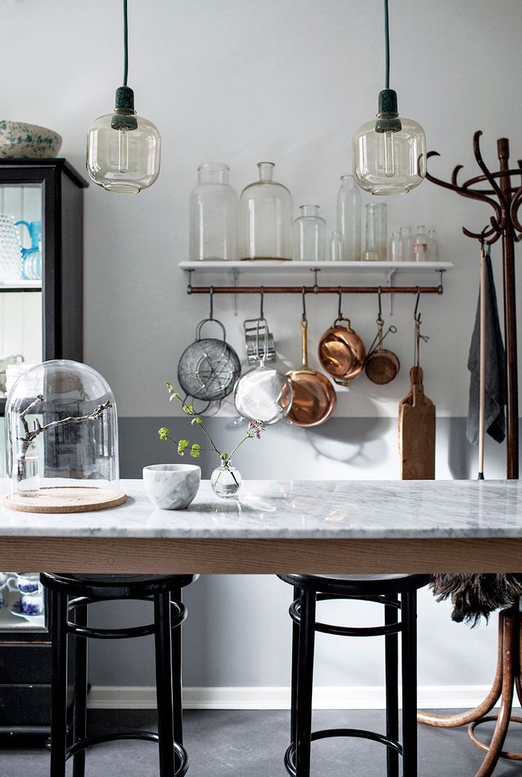 Modern kitchen with hanging copper pots and metal utensils, featuring glass pendant lights and a marble countertop with simple greenery in glass vases