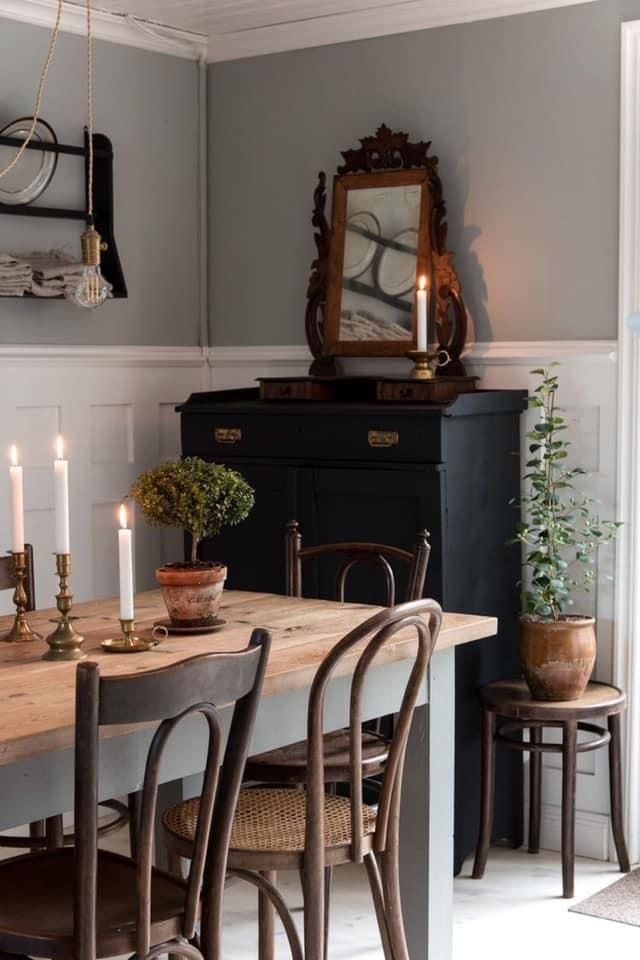 A rustic Scandinavian dining space with a wooden table and vintage chairs. Candles and simple potted plants add warmth, while a black cabinet with an antique mirror enhances the cozy, minimalist atmosphere