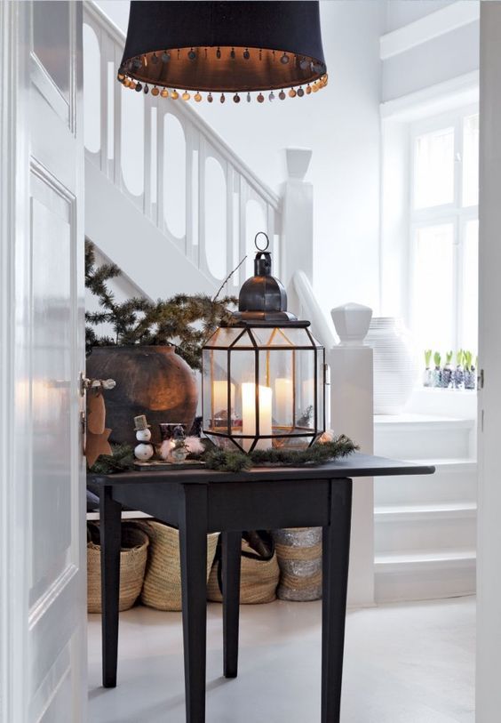 A black accent table with a large lantern centerpiece decorated with candles and pine branches. The table is positioned in a white entryway with a modern black chandelier above. The clean, minimalist surroundings contrast with the festive decor on the table, creating a cozy atmosphere.