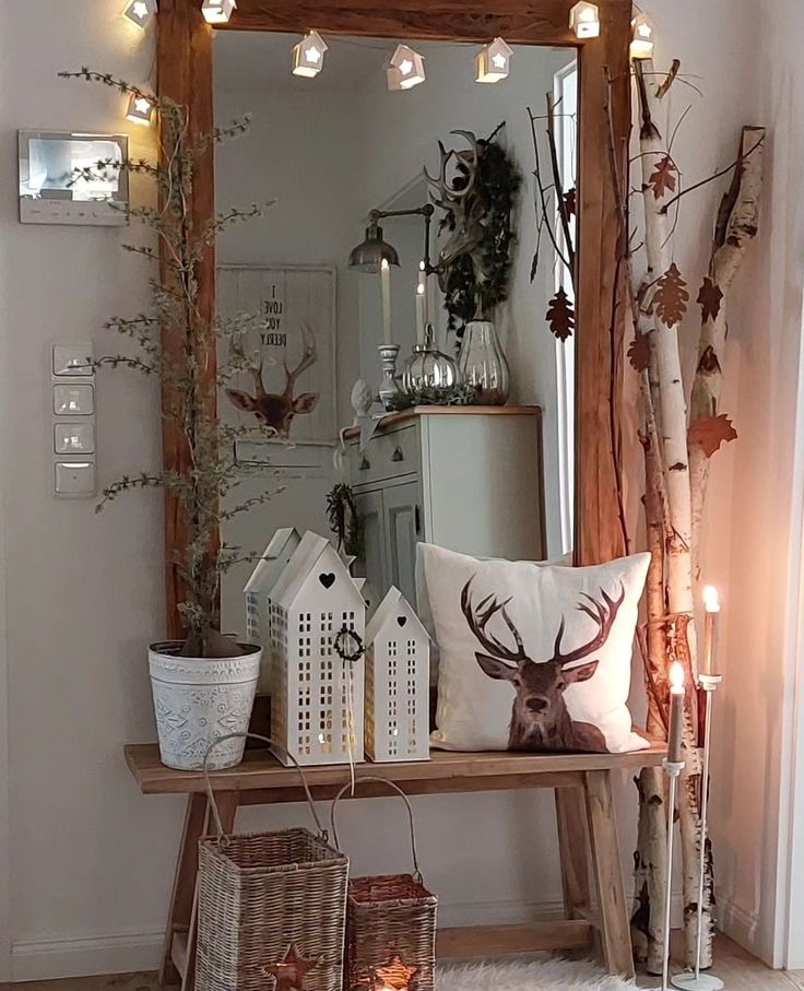 An entryway with natural and rustic holiday decor, featuring a large mirror framed with small house-shaped string lights, a reindeer print pillow, and minimalistic winter accents. Birch branches, wicker baskets, and small, white ceramic houses add a cozy, woodland-inspired feel to the space.