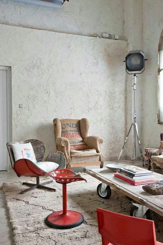 An eclectic living room with rough white plaster walls, mismatched vintage seating, and a mix of industrial and rustic furniture. A red metal chair stands out against the muted tones, along with a large industrial floor lamp and a reclaimed wood coffee table on a textured rug.