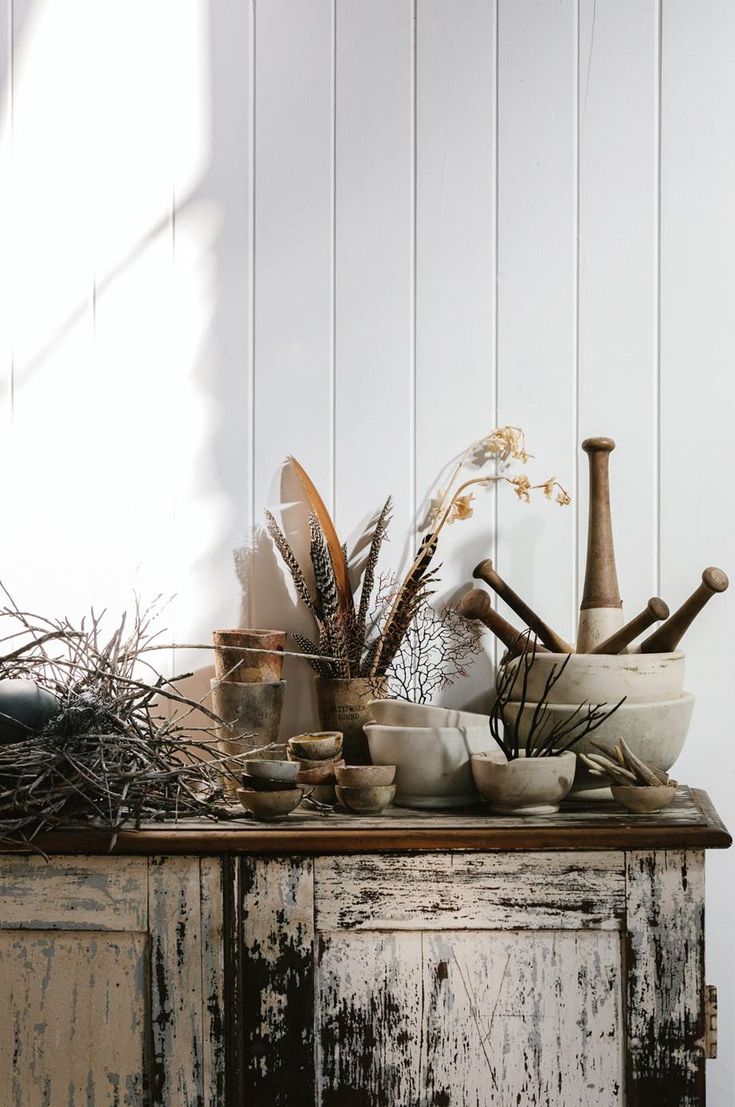 Rustic vignette on a weathered wooden sideboard, displaying dried branches, mortar and pestles, feathers, and small earthenware bowls. The setup captures a minimalist yet earthy, natural aesthetic with a white wood-paneled backdrop.