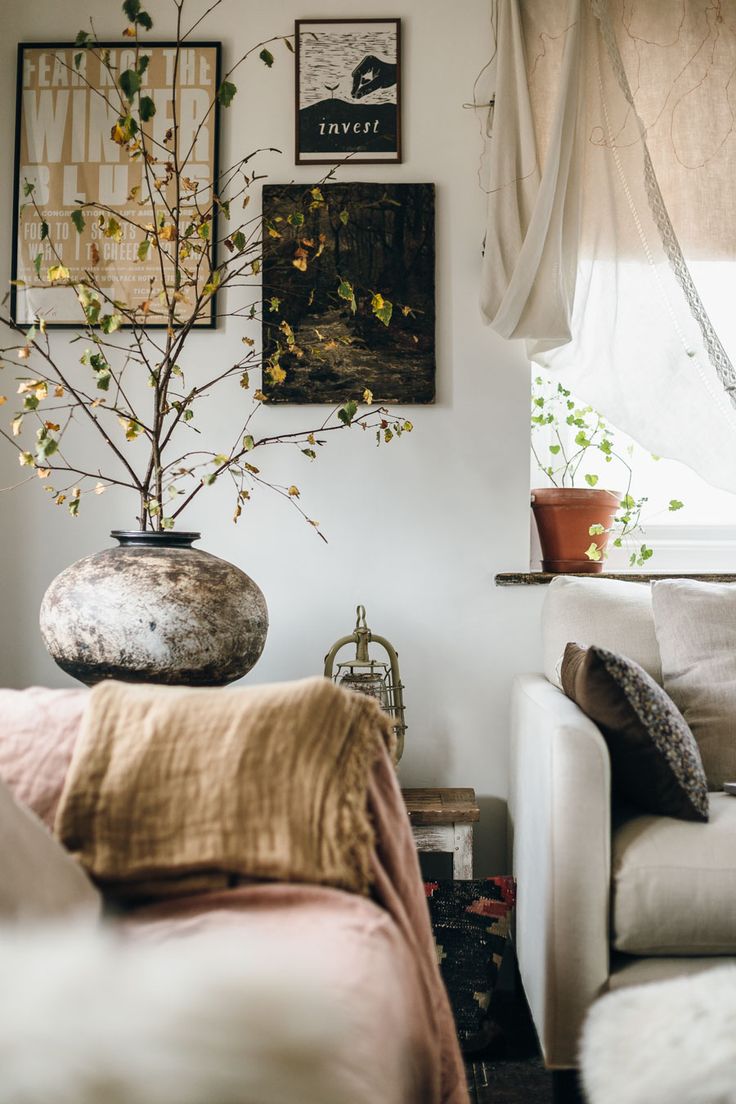Cozy living room styled with natural elements, featuring a large rustic vase holding tree branches, warm earthy tones in furniture and textiles, and potted plants on the windowsill.