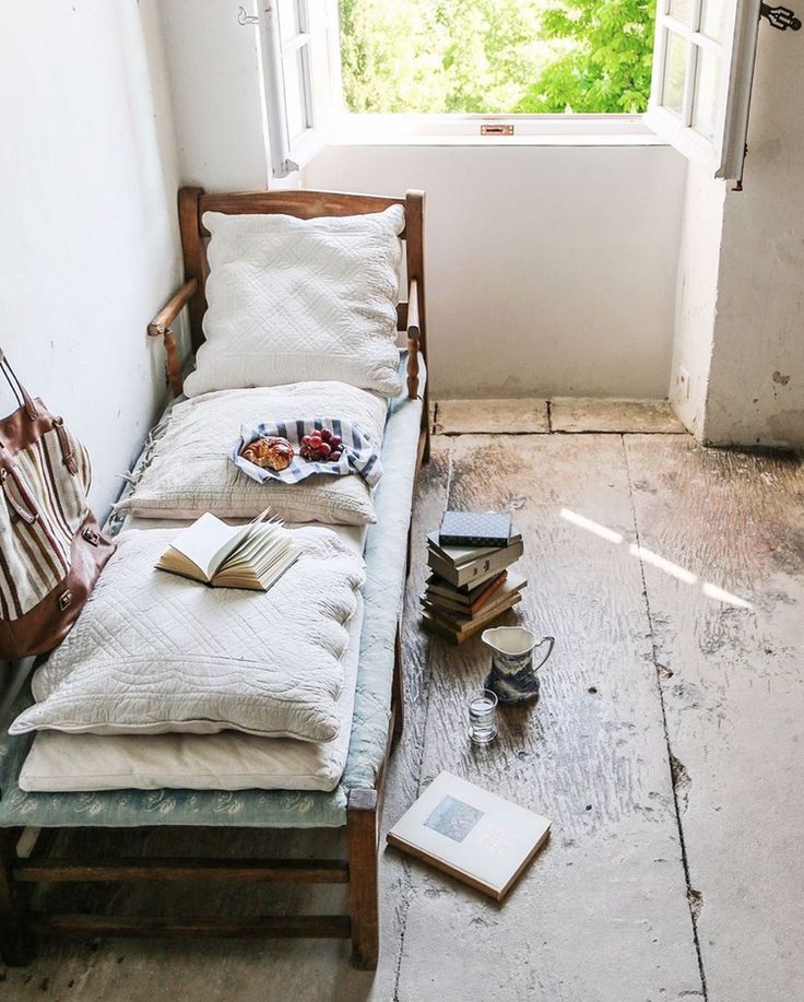 Charming reading nook by an open window, showcasing a rustic daybed adorned with white, quilted pillows. A stack of books, a plate of snacks, and a vintage pitcher complete the relaxed, serene atmosphere.