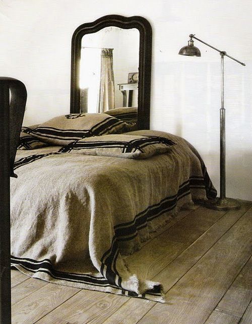 Cozy bedroom with a rustic, vintage vibe featuring a neutral-toned bed covered with a textured burlap throw blanket. A black-framed mirror stands behind the bed, and a sleek industrial floor lamp adds a modern touch to the space.