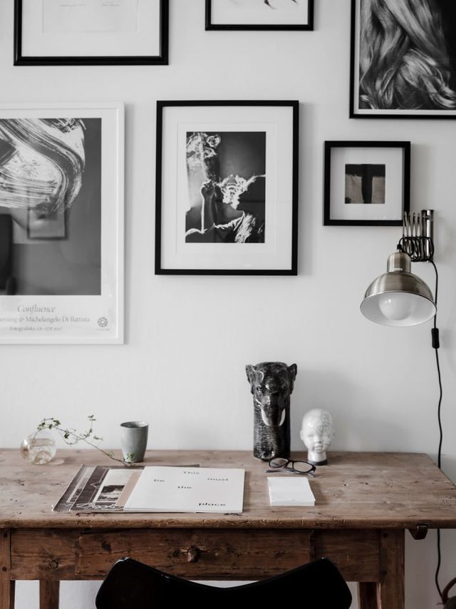 A black-and-white gallery wall with abstract and portrait art above a rustic wooden desk. The workspace is minimalist yet stylish, with eclectic decor items like a black panther statue and a small white bust, adding a mysterious, modern feel to the space.