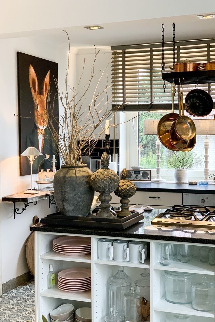 A bright dining area with a kitchen island decorated with a large ceramic vase, sculptural accents, and copper pots hanging overhead, blending modern and rustic styles with playful artwork in the background.