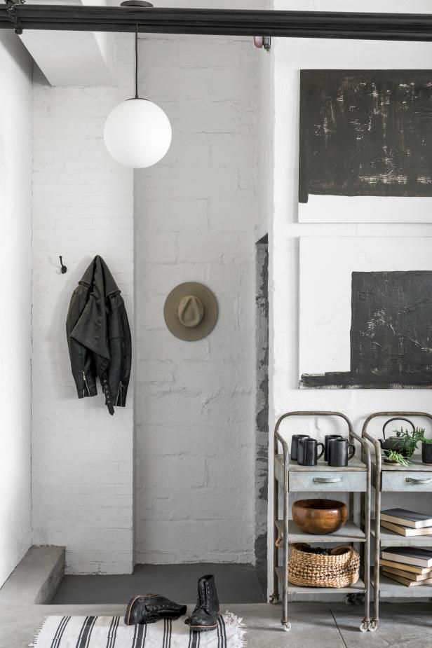 Entryway featuring a galvanized metal cart decorated with black mugs, wooden bowls, books, and a plant, accompanied by a black leather jacket and hat hung on the white brick wall, creating a modern industrial vibe.