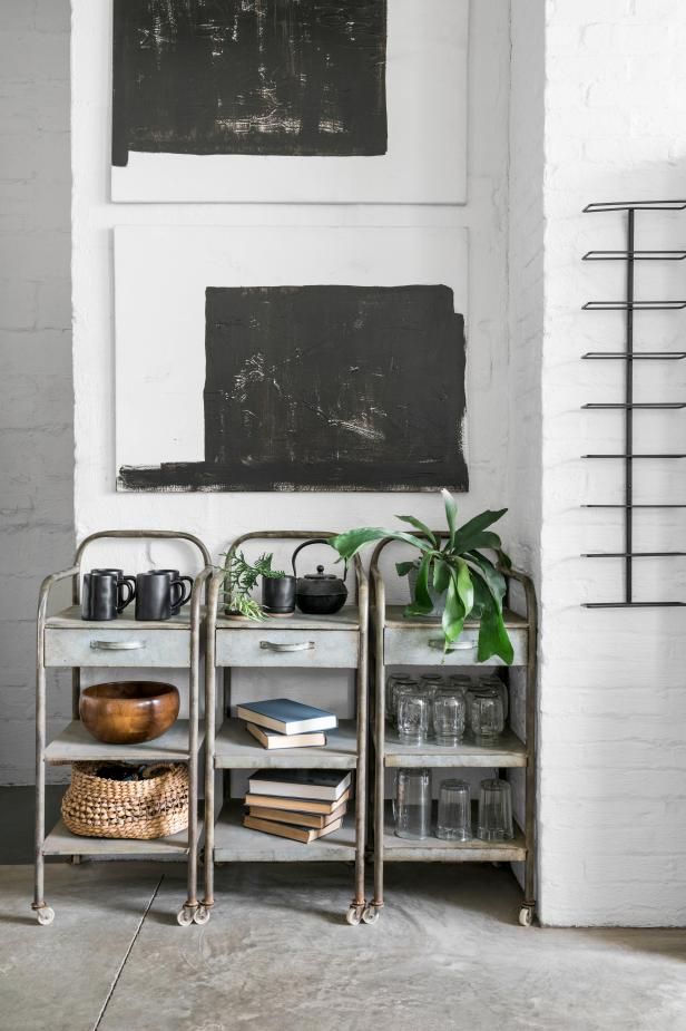 Three galvanized metal storage carts used as stylish shelving, decorated with black mugs, plants, books, and glass jars, placed in a white brick-walled space with modern abstract black artwork.