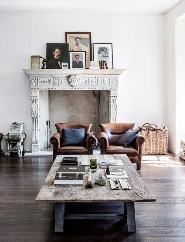 A living room featuring a rustic wooden coffee table, two leather armchairs with throw pillows, and a decorated mantel displaying vintage-style portrait art in various frames