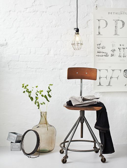 A minimalist industrial setting featuring a metal stool with a wooden seat, a hanging light bulb fixture, a large glass vase with fresh greenery, and a white brick wall adorned with a typography poster. The overall look is clean and modern with a rustic touch.