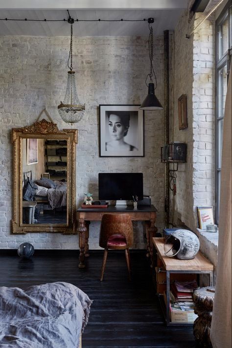 An industrial-style room with a white brick wall, a vintage wooden desk, a large gold-framed mirror, and a striking black-and-white portrait as the focal point.