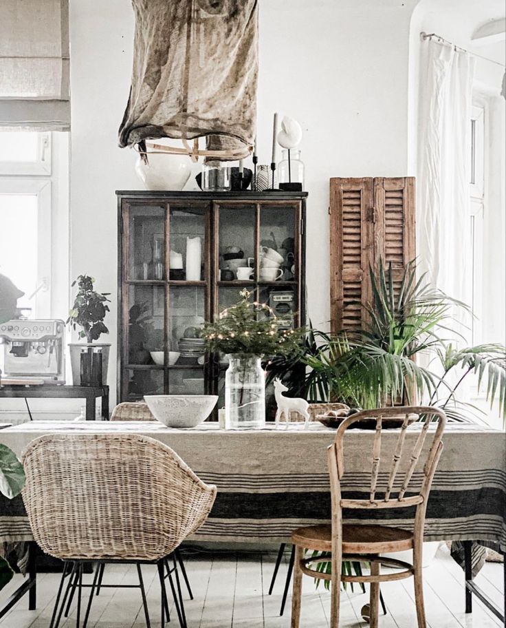 A dining room styled with mismatched rattan and wooden chairs, a striped tablecloth, lush greenery, a vintage cabinet filled with crockery, and rustic decor elements, creating an eclectic and inviting space.