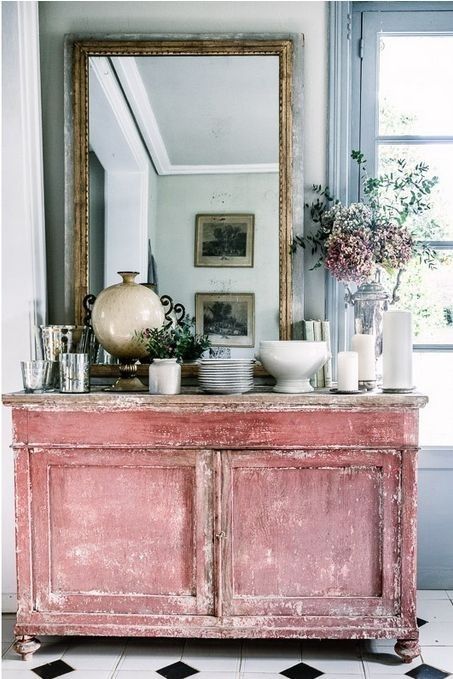 Vintage pink distressed cabinet topped with elegant glassware, a ceramic vase, candles, and floral arrangements, paired with a large gold-framed mirror reflecting a cozy interior setting.