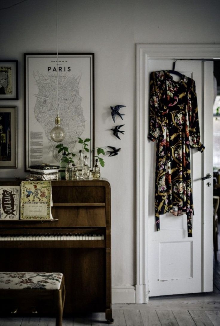 A rustic upright piano styled with vintage sheet music, glass bottles, and greenery. The setting includes a Paris map print, swallows on the wall, and a colorful patterned dress hanging nearby, creating a moody eclectic vibe.