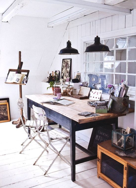 A cozy and artistic home office with a gallery wall showcasing eclectic framed artwork and portraits. The space includes a metal shelving unit filled with creative supplies, wooden floors, and a glass-topped desk with mid-century modern chairs.