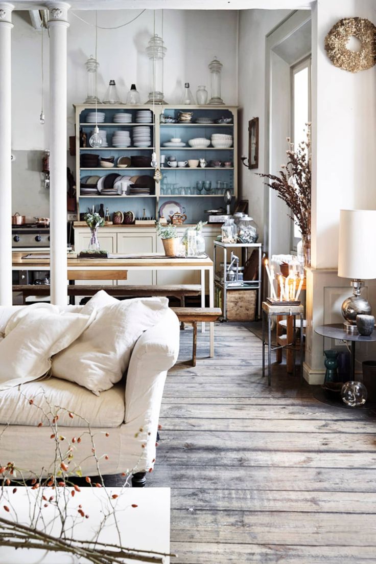 A bright and airy open-concept kitchen and dining space with a vintage aesthetic. A distressed wooden floor, open shelving filled with neatly arranged dishware, and natural elements like dried flowers and glass jars create a charming shabby chic atmosphere.