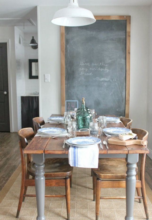 A rustic dining room with a wooden farmhouse table featuring gray-painted legs, surrounded by vintage wooden chairs. A large chalkboard with a wooden frame serves as a decorative focal point, adding a cozy and personal touch to the space.