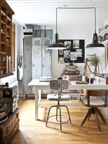 An eclectic workspace featuring a vintage industrial style with distressed lockers, wooden desks, and black pendant lighting. The space is decorated with vintage suitcases, framed art, and rustic elements that add character.