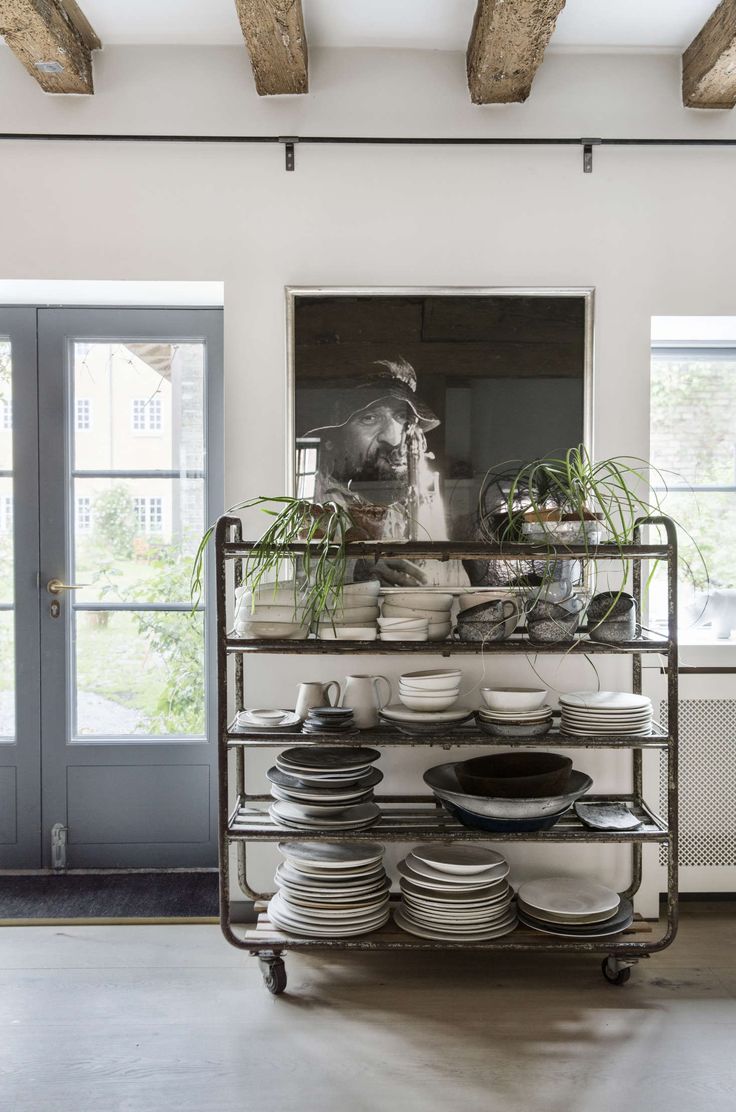 Industrial-style open shelving filled with stacked plates, bowls, and kitchen essentials, complemented by hanging plants and rustic wooden beams in a bright, airy kitchen space.