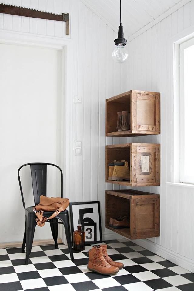 Minimalist entryway with black and white checkered flooring, rustic wooden crates mounted on the wall, a black metal chair, and simple decor creating a clean, graphic look.