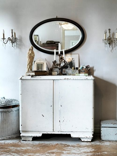 A vintage white distressed cabinet with an oval mirror above it, surrounded by antique wall sconces, decorative candles, and a rustic statue. The setting exudes a charming, eclectic style.